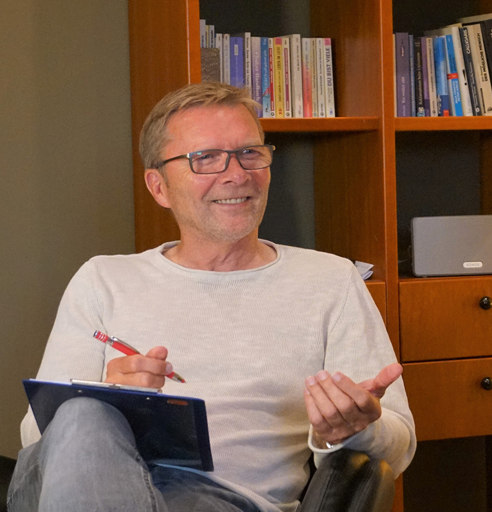 Erich Thoben, der lässig in einem Stuhl sitzt. Er trägt ein weißes Langarmshirt und graue Hosen. In seiner linken Hand hält er ein geöffnetes Notizbuch und einen Stift, während seine rechte Hand eine erklärende Geste macht. Im Hintergrund ist ein Bücherregal mit einer Reihe von Büchern und ein Laptop auf einem Schrank zu sehen